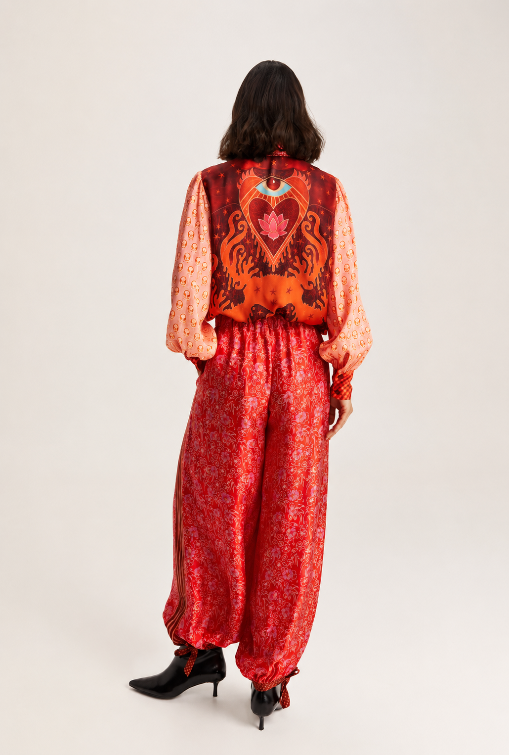 A model viewed from the back wearing a vibrant red and orange patterned jumpsuit with a large heart design on the back, paired with black ankle boots, against a plain light gray studio background.