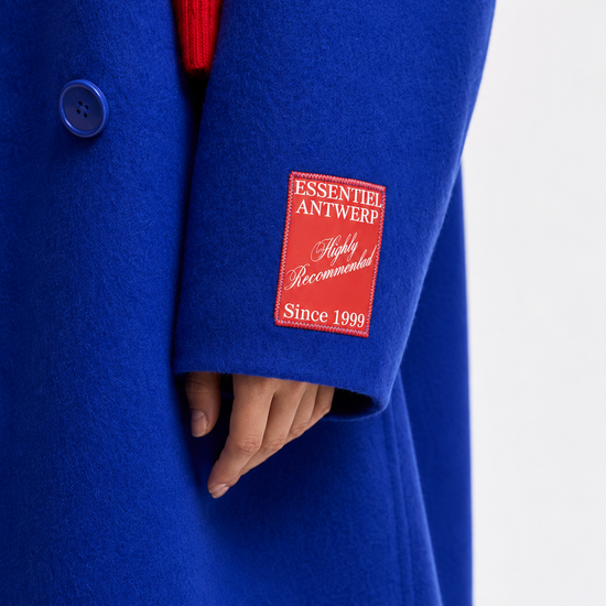 A close-up of a bright royal blue wool coat worn over a red ribbed sweater, featuring a red brand patch on the sleeve that reads 'ESSENTEI ANTWERP Highly Recommended Since 1998'.