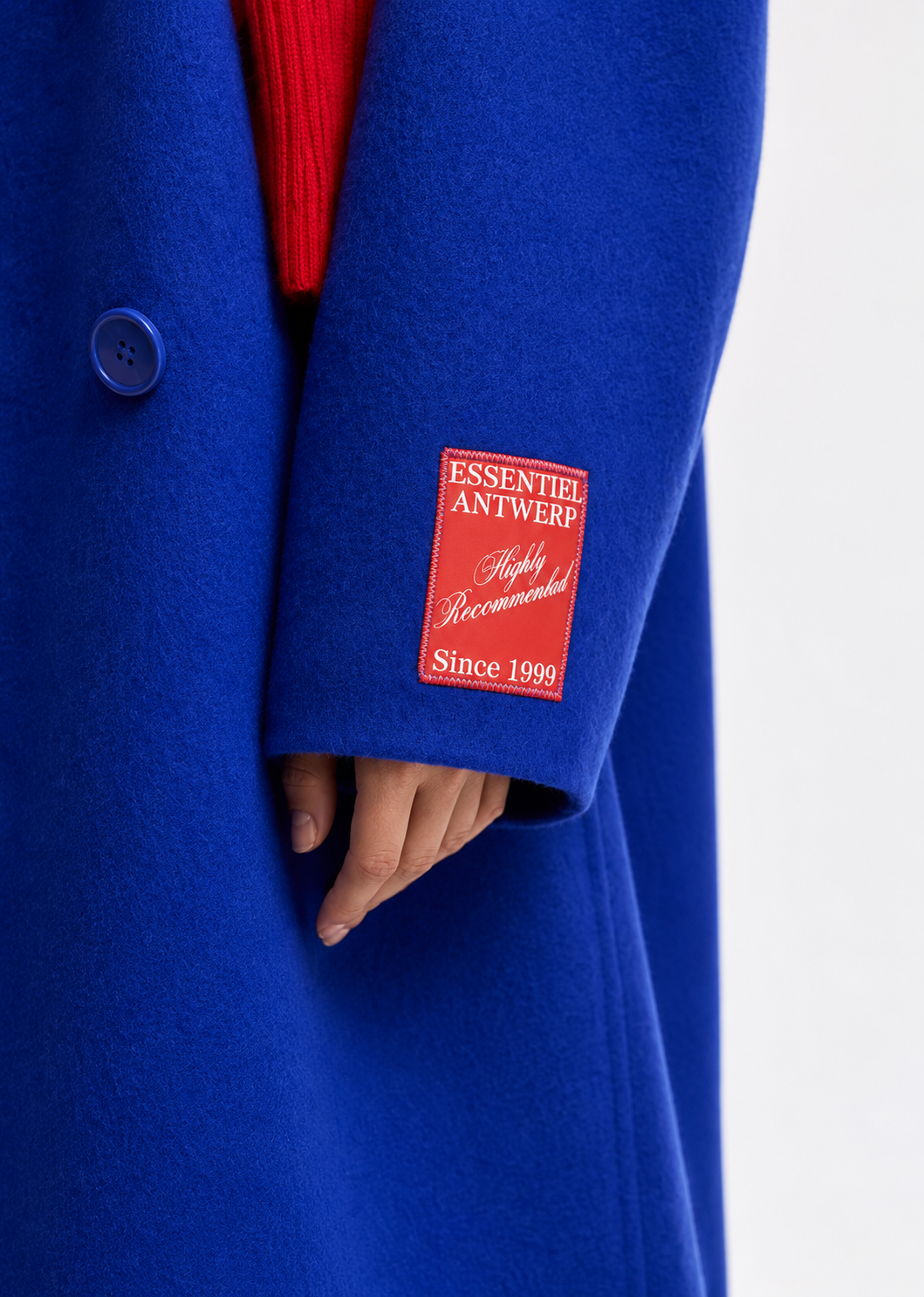 A close-up of a bright royal blue wool coat worn over a red ribbed sweater, featuring a red brand patch on the sleeve that reads 'ESSENTEI ANTWERP Highly Recommended Since 1998'.