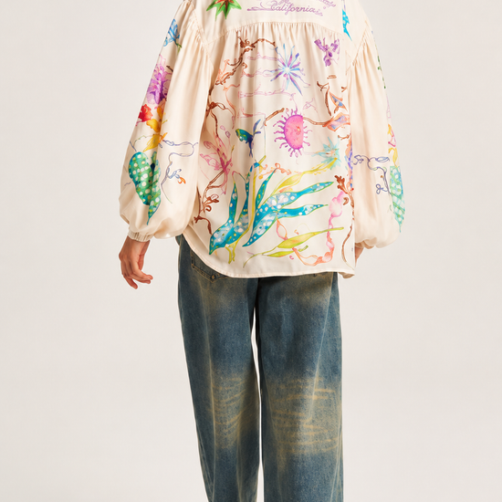 A woman shown from the back wearing a cream-colored, long-sleeved blouse with colorful floral and botanical illustrations, paired with wide-leg blue jeans and black heels, against a light gray studio background.