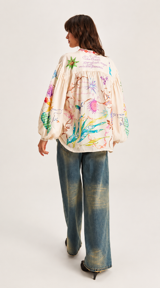 A woman shown from the back wearing a cream-colored, long-sleeved blouse with colorful floral and botanical illustrations, paired with wide-leg blue jeans and black heels, against a light gray studio background.