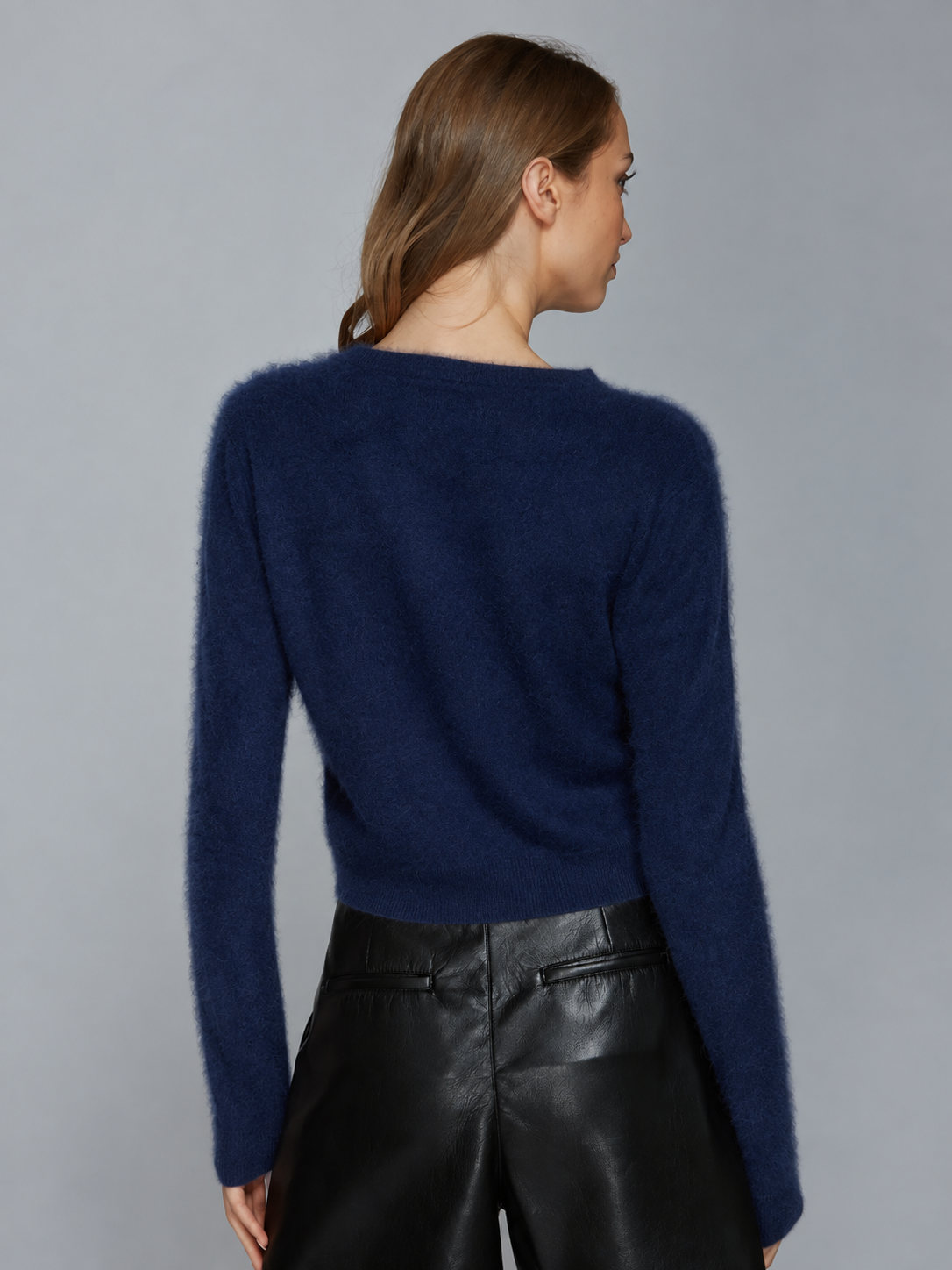 A woman shown from the back wearing a fuzzy navy blue long-sleeve sweater and black leather pants against a plain gray studio background.