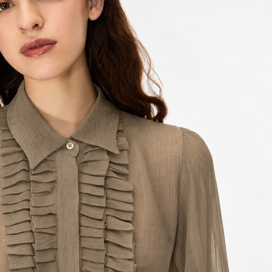 A close-up of a model wearing a taupe brown ruffled blouse with a pointed collar and a single button, set against a plain white background.