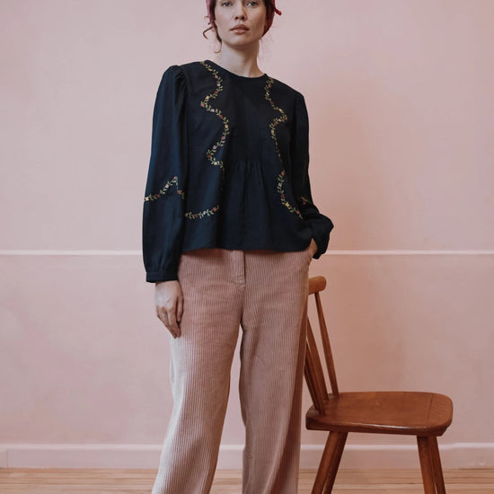 A woman stands in a room with a pink wall and wooden floor, wearing a black long-sleeve blouse with gold embroidered wavy trim and light pink ribbed trousers. A wooden chair is beside her, and she wears a pink headscarf.