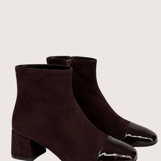 A pair of dark brown suede ankle boots with glossy patent leather cap toes and low block heels, shown against a light beige background.