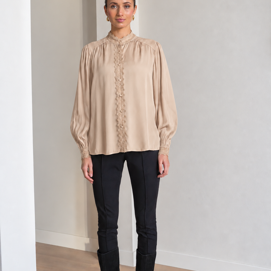 A woman modeling a beige long-sleeve blouse with a high gathered neckline and lace-up front, paired with black pants and black boots, standing in a minimalist room with light gray walls and wood floors.