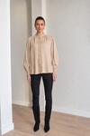 A woman modeling a beige long-sleeve blouse with a high gathered neckline and lace-up front, paired with black pants and black boots, standing in a minimalist room with light gray walls and wood floors.