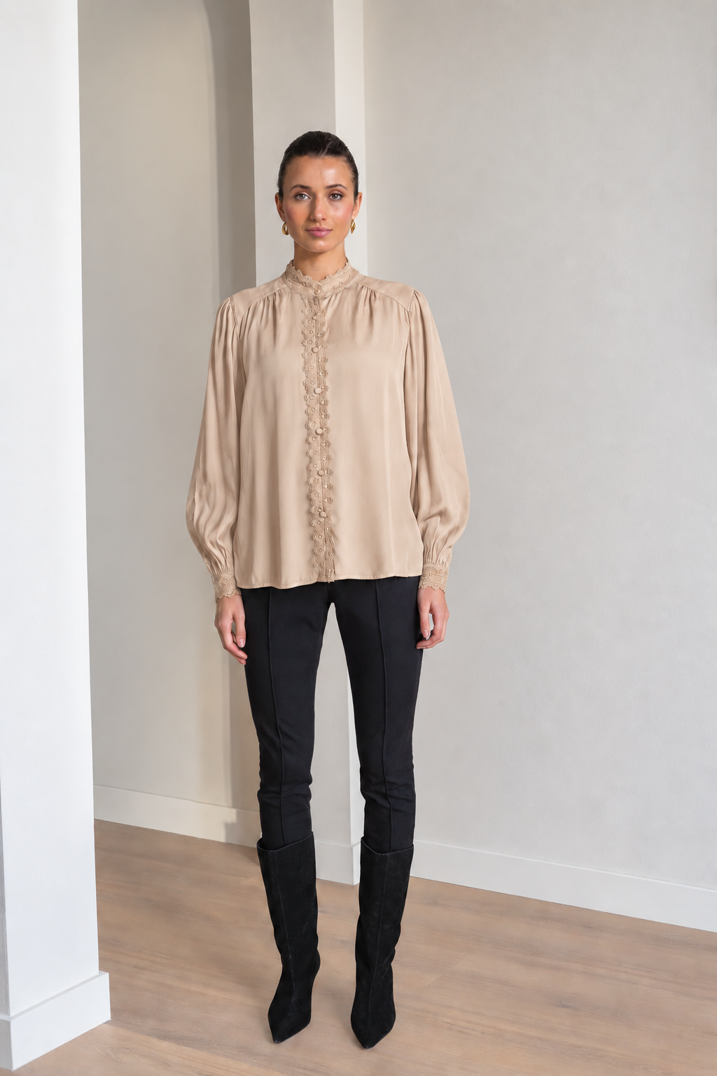 A woman modeling a beige long-sleeve blouse with a high gathered neckline and lace-up front, paired with black pants and black boots, standing in a minimalist room with light gray walls and wood floors.