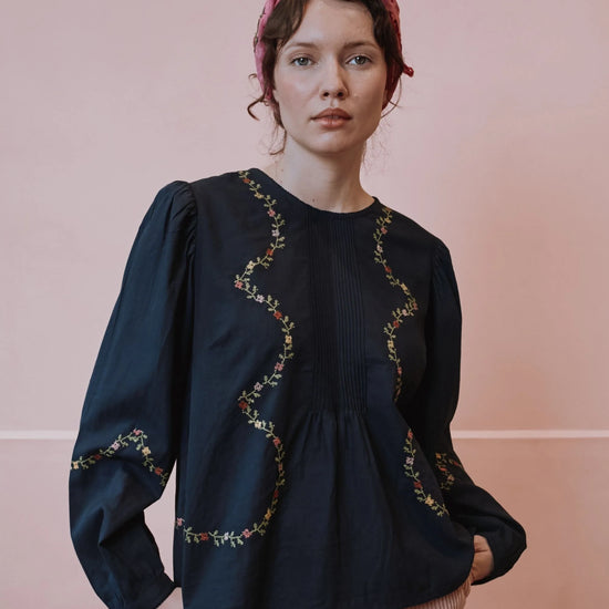A woman wearing a dark navy blue long-sleeve blouse with wavy floral embroidery, paired with pink striped trousers and a pink headscarf, standing against a solid pink background.
