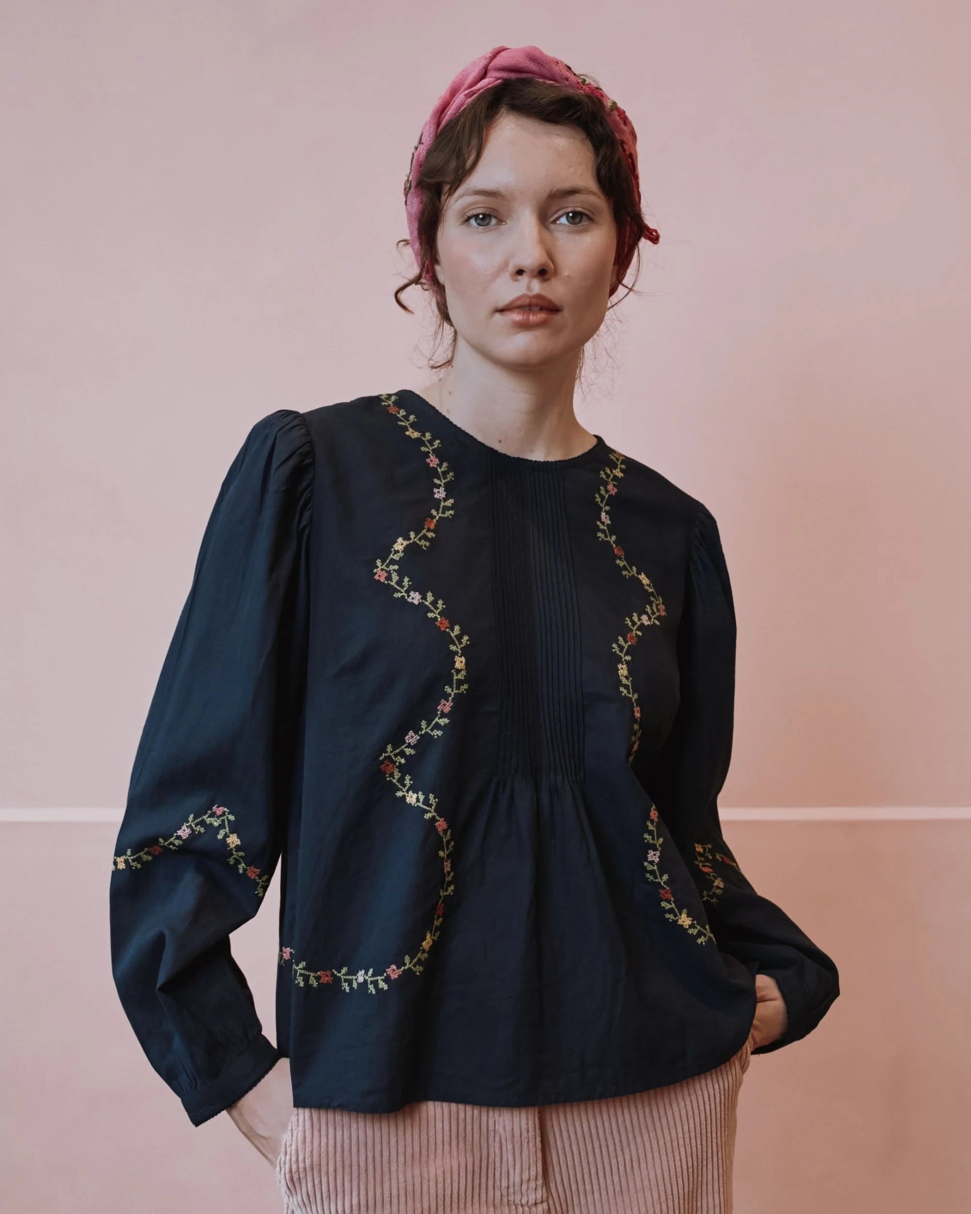 A woman wearing a dark navy blue long-sleeve blouse with wavy floral embroidery, paired with pink striped trousers and a pink headscarf, standing against a solid pink background.