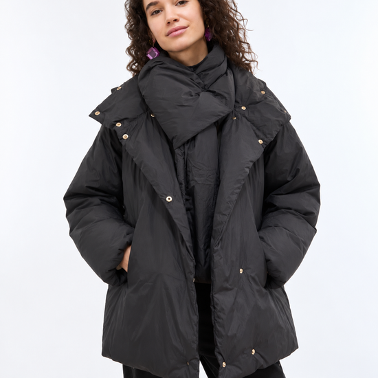 A woman modeling a black oversized puffer jacket with a matching oversized scarf, styled with black pants on a light gray studio background.