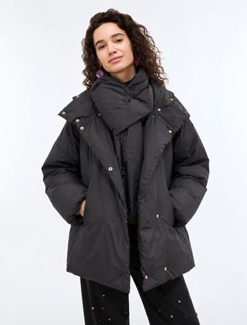 A woman modeling a black oversized puffer jacket with a matching oversized scarf, styled with black pants on a light gray studio background.
