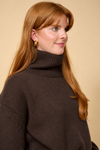 A woman with long red hair wearing a dark brown ribbed turtleneck sweater and small gold hoop earrings, photographed against a plain beige background.