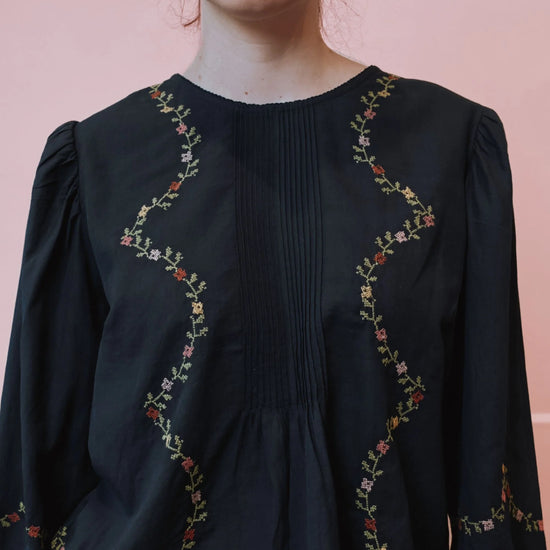 A close-up of a woman wearing a black long-sleeve blouse with vertical pleating and wavy floral embroidery in red, green, and yellow along the neckline and sleeves, set against a solid pink background.