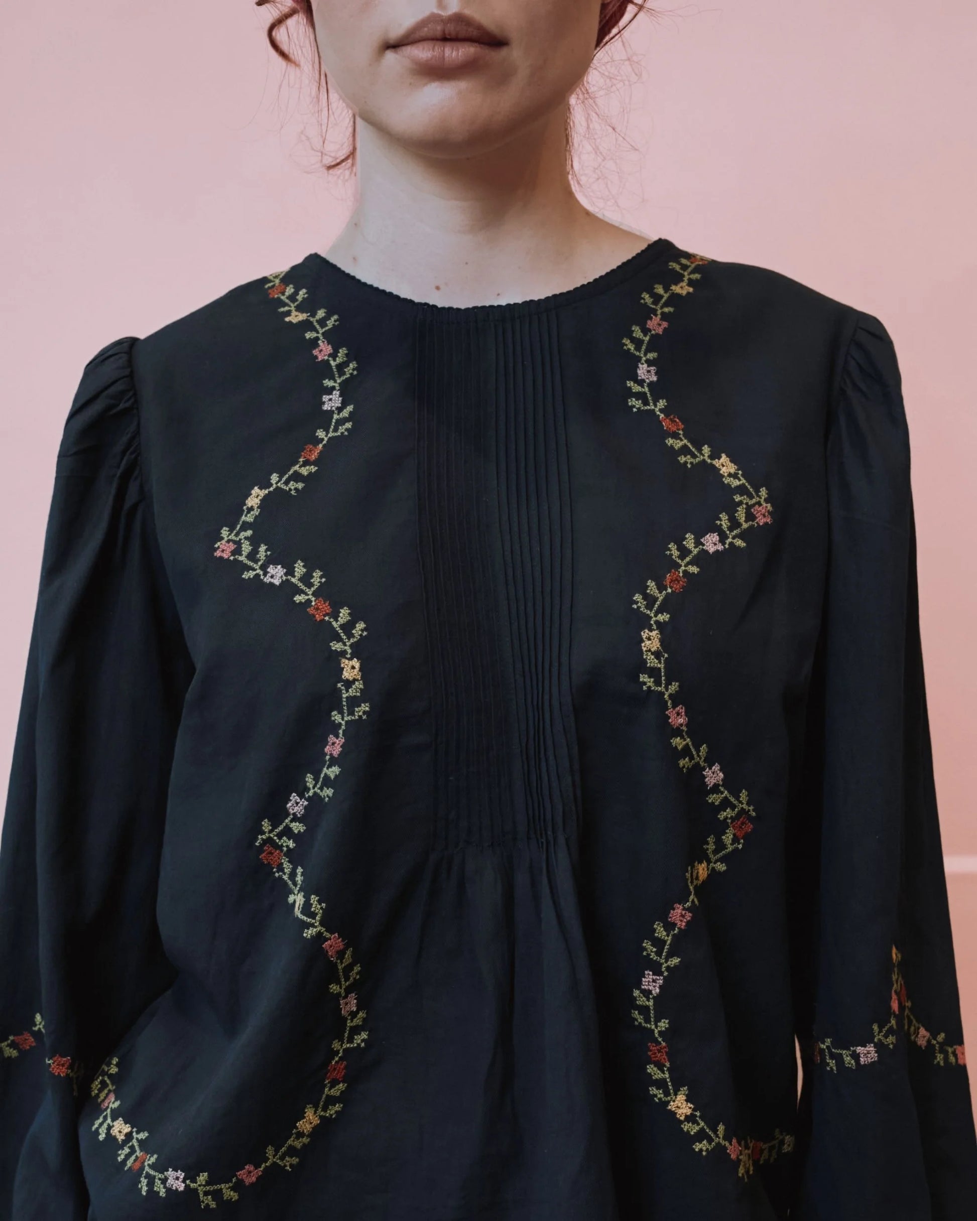 A close-up of a woman wearing a black long-sleeve blouse with vertical pleating and wavy floral embroidery in red, green, and yellow along the neckline and sleeves, set against a solid pink background.