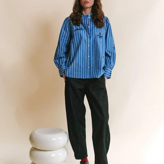 A model stands in a studio wearing a blue and white vertically striped button-up shirt, black trousers, and black leather shoes, next to a stacked white ceramic stool against a beige background.
