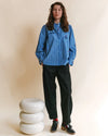 A model stands in a studio wearing a blue and white vertically striped button-up shirt, black trousers, and black leather shoes, next to a stacked white ceramic stool against a beige background.