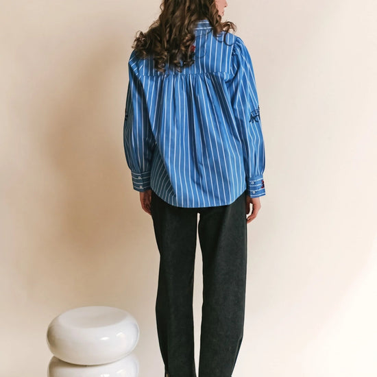 A woman shown from the back wearing a blue and white vertically striped long-sleeve shirt, dark charcoal pants, and sneakers, standing in front of a beige studio background with a small white stacked stool at her left.