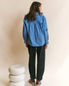 A woman shown from the back wearing a blue and white vertically striped long-sleeve shirt, dark charcoal pants, and sneakers, standing in front of a beige studio background with a small white stacked stool at her left.