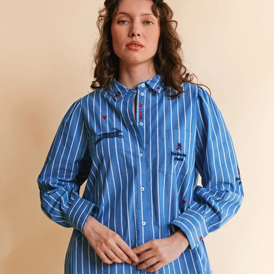A woman wearing a blue and white vertically striped button-up shirt with embroidered details, standing against a solid beige background.