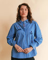 A woman wearing a blue and white vertically striped button-up shirt with embroidered details, standing against a solid beige background.