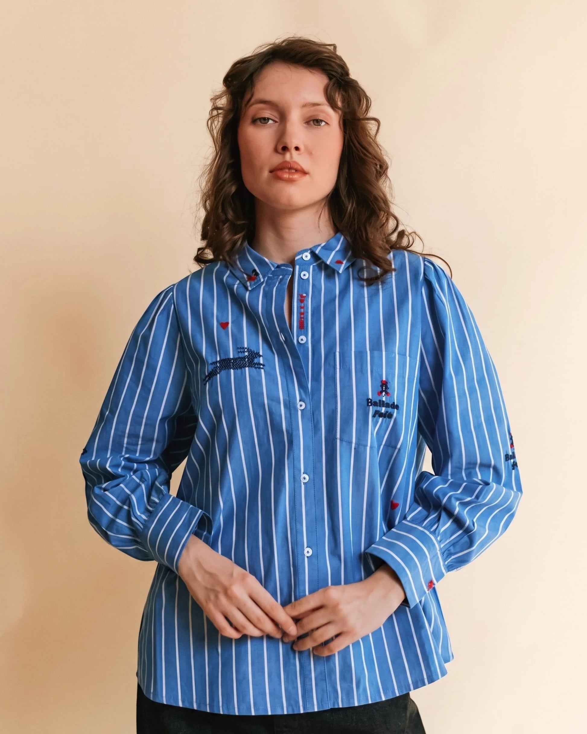 A woman wearing a blue and white vertically striped button-up shirt with embroidered details, standing against a solid beige background.