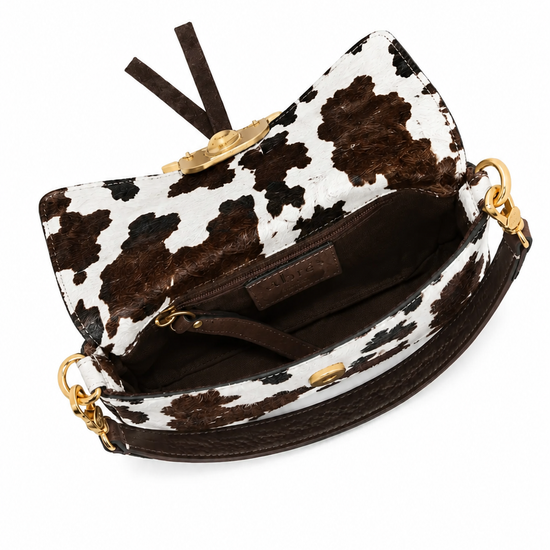 A cow-print clutch handbag with brown faux-leather trim, gold-tone hardware, and a dark brown interior, shown from an overhead angle on a white background.