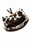 A cow-print clutch handbag with brown faux-leather trim, gold-tone hardware, and a dark brown interior, shown from an overhead angle on a white background.