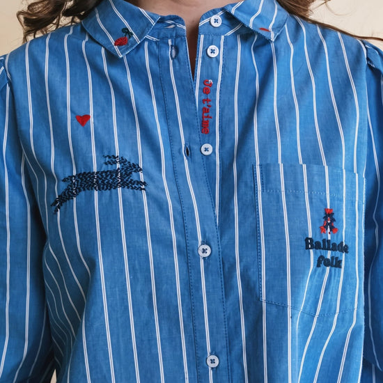 A close-up of a person wearing a blue button-up shirt with white vertical pinstripes. The shirt features embroidered details, including a crocodile motif on the chest, a small red heart, and a pocket with a Balenciaga logo.