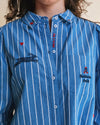 A close-up of a person wearing a blue button-up shirt with white vertical pinstripes. The shirt features embroidered details, including a crocodile motif on the chest, a small red heart, and a pocket with a Balenciaga logo.