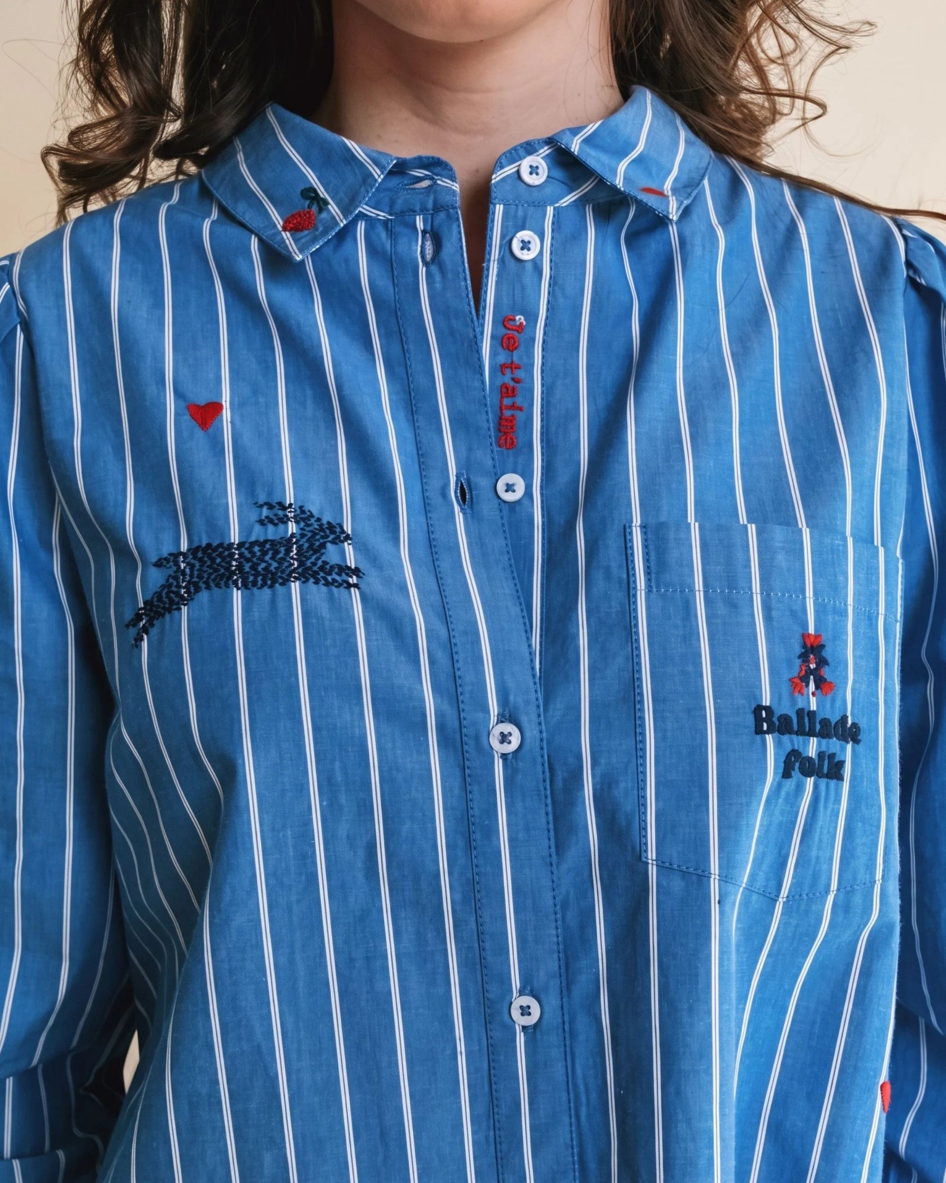 A close-up of a person wearing a blue button-up shirt with white vertical pinstripes. The shirt features embroidered details, including a crocodile motif on the chest, a small red heart, and a pocket with a Balenciaga logo.