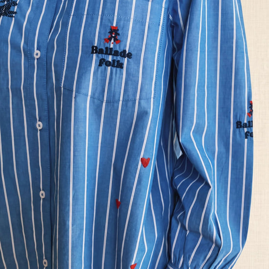 A close-up of a blue and white vertically striped long-sleeve button-up shirt with 'Ballade Folk' embroidered on the chest pocket.