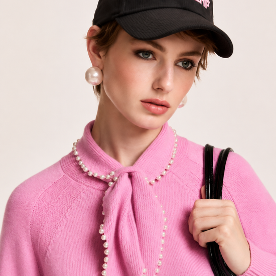 A model wearing a pink knit sweater with a pearl-trimmed bow collar, a black baseball cap with pink 'ESSENTIAL ANTWERP' embroidery, and pearl earrings, holding a black shoulder bag against a light beige background.