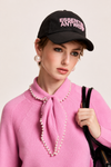 A model wearing a pink knit sweater with a pearl-trimmed bow collar, a black baseball cap with pink 'ESSENTIAL ANTWERP' embroidery, and pearl earrings, holding a black shoulder bag against a light beige background.