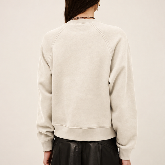 A woman shown from the back wearing a cream-colored crewneck sweatshirt and a black pleated mini skirt, set against a plain light beige studio background.