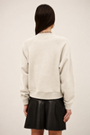 A woman shown from the back wearing a cream-colored crewneck sweatshirt and a black pleated mini skirt, set against a plain light beige studio background.