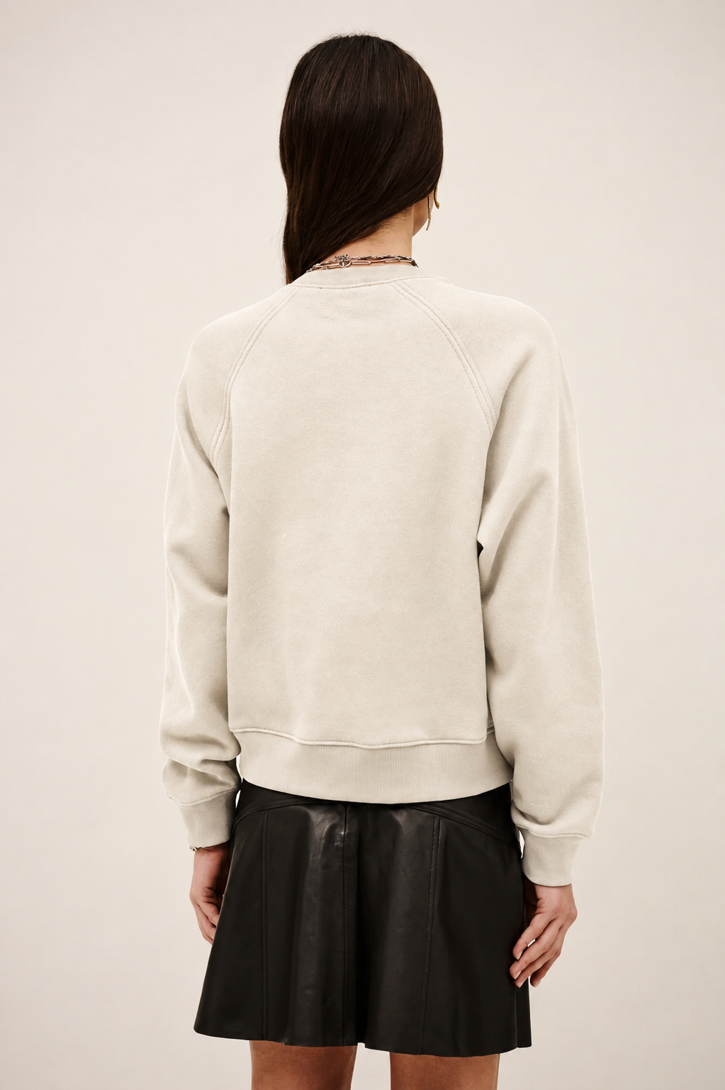 A woman shown from the back wearing a cream-colored crewneck sweatshirt and a black pleated mini skirt, set against a plain light beige studio background.