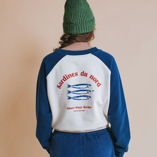 A person shown from the back wearing a raglan-style sweatshirt with blue sleeves and a white back panel featuring red text reading 'sardines du nord' and blue fish graphics, paired with blue corduroy pants and a green knit beanie against a plain beige background.
