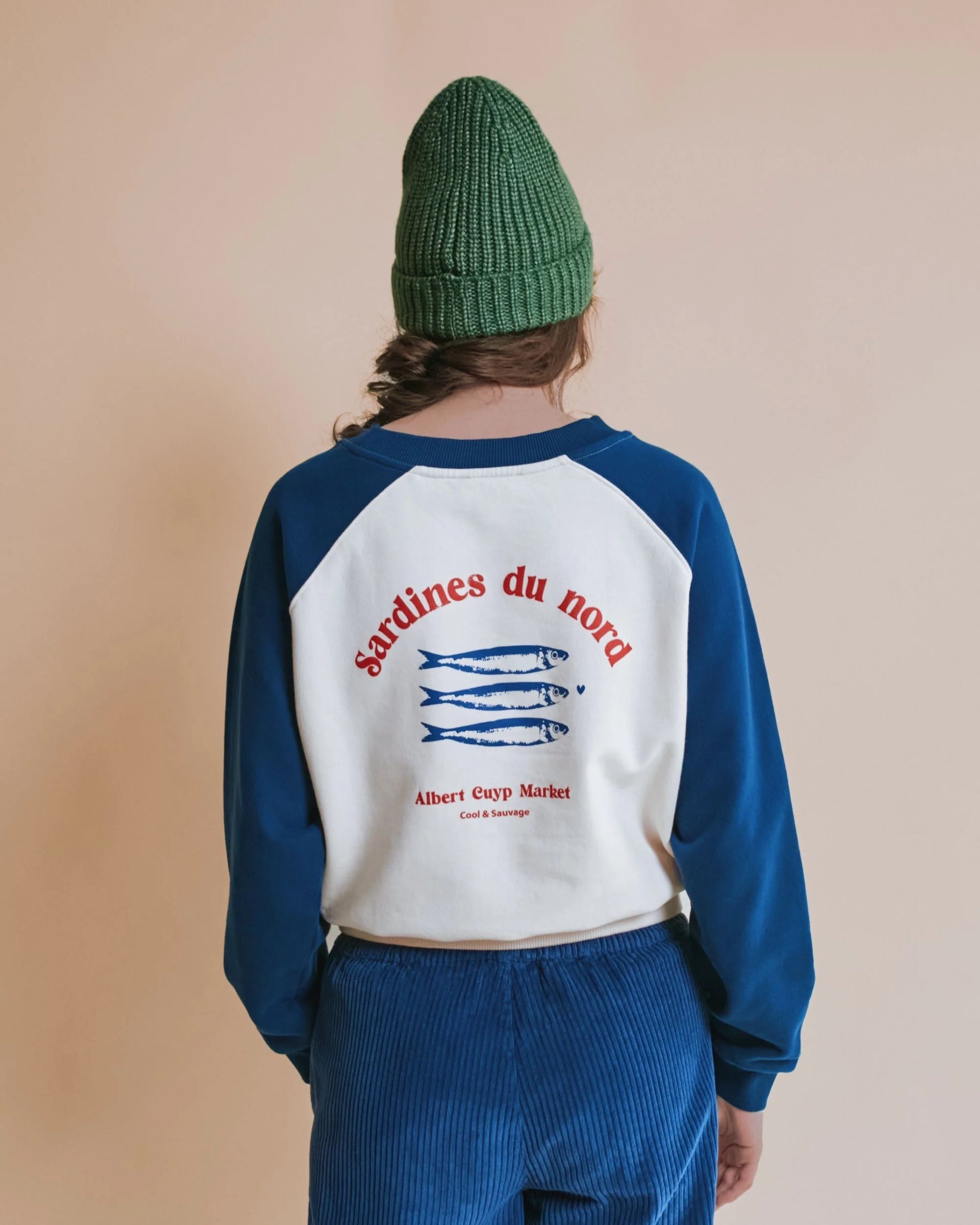 A person shown from the back wearing a raglan-style sweatshirt with blue sleeves and a white back panel featuring red text reading 'sardines du nord' and blue fish graphics, paired with blue corduroy pants and a green knit beanie against a plain beige background.