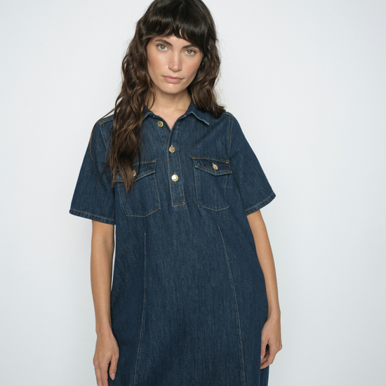 A woman modeling a dark blue denim short-sleeve shirt dress with button details and chest pockets, set against a plain light gray background.