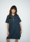 A woman modeling a dark blue denim short-sleeve shirt dress with button details and chest pockets, set against a plain light gray background.