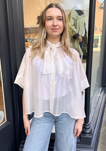 Woman wearing a white blouse with a bow tie in front with blue jeans.