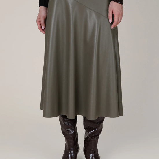 A model is shown from the waist down wearing a mid-length, asymmetrical wrap skirt in a dark olive green faux leather, paired with dark brown boots and a black long-sleeve top against a plain beige studio background.