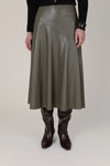 A model is shown from the waist down wearing a mid-length, asymmetrical wrap skirt in a dark olive green faux leather, paired with dark brown boots and a black long-sleeve top against a plain beige studio background.
