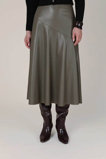 A model is shown from the waist down wearing a mid-length, asymmetrical wrap skirt in a dark olive green faux leather, paired with dark brown boots and a black long-sleeve top against a plain beige studio background.
