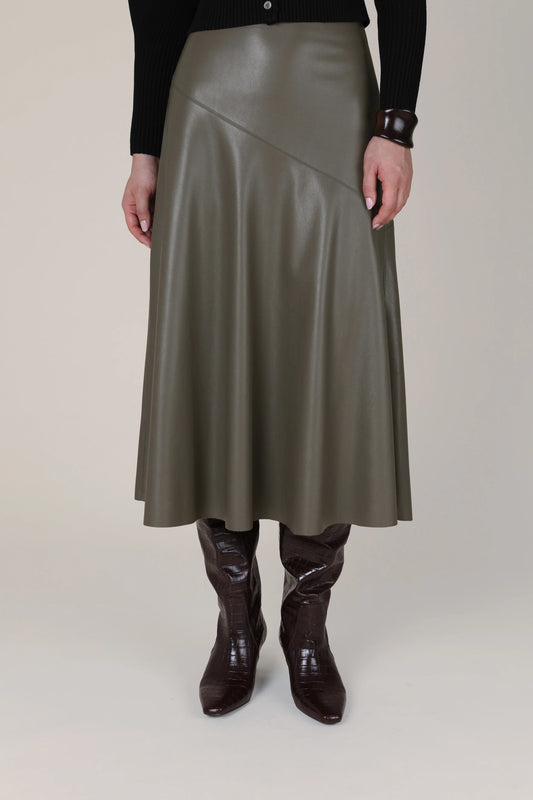 A model is shown from the waist down wearing a mid-length, asymmetrical wrap skirt in a dark olive green faux leather, paired with dark brown boots and a black long-sleeve top against a plain beige studio background.