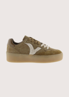 A low-top platform sneaker in olive green suede and white faux fur, featuring a thick tan sole and white laces, shown from a side profile on a light gray background.