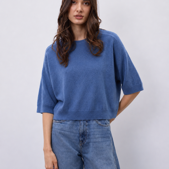 A woman modeling a blue short-sleeve knit top with wide legs, paired with blue jeans, against a light gray studio background.
