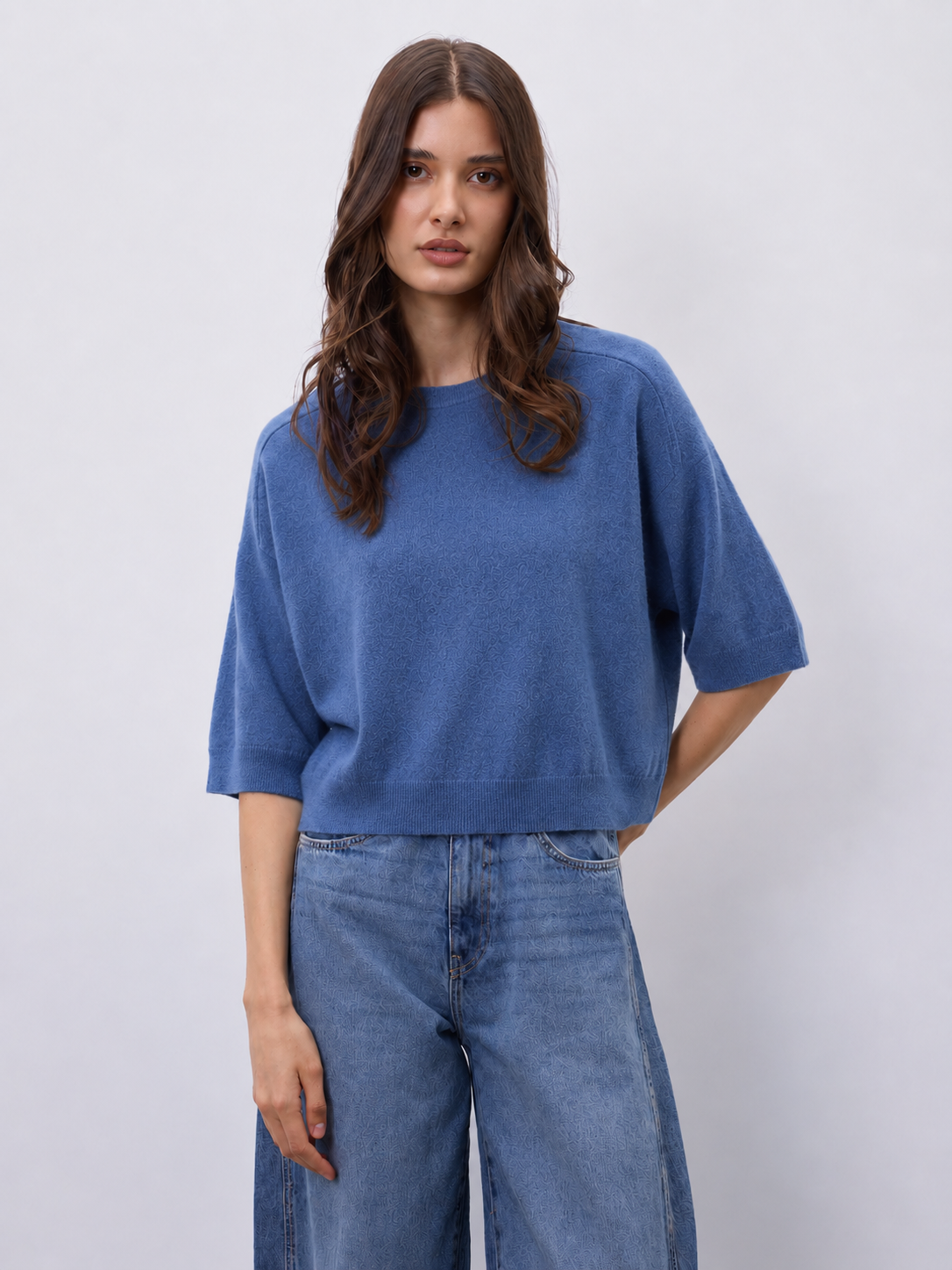 A woman modeling a blue short-sleeve knit top with wide legs, paired with blue jeans, against a light gray studio background.