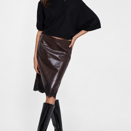 A model wearing a black off-the-shoulder knit top, a shiny dark brown faux-leather midi skirt, and black knee-high boots, posed against a light gray studio background.